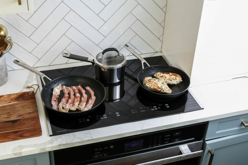 Bacon being cooked in a pan, eggs being boiled in a pot, and chicken being cooked on the burner zones of a Sharp Induction Cooktop SCH3042GB