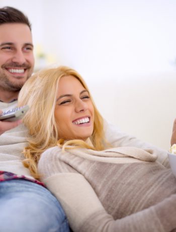 Happy young couple lying on the sofa at home with popcorn watching TV. They are laughing and watching a movie or television.