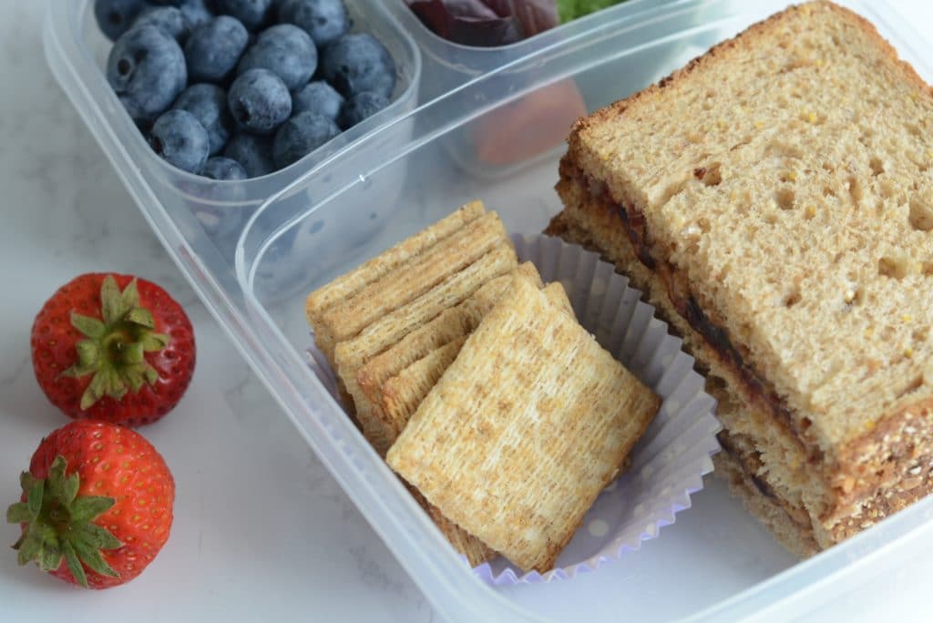 Sandwhich in a containerwith crackers, blueberries, and strawberries.
