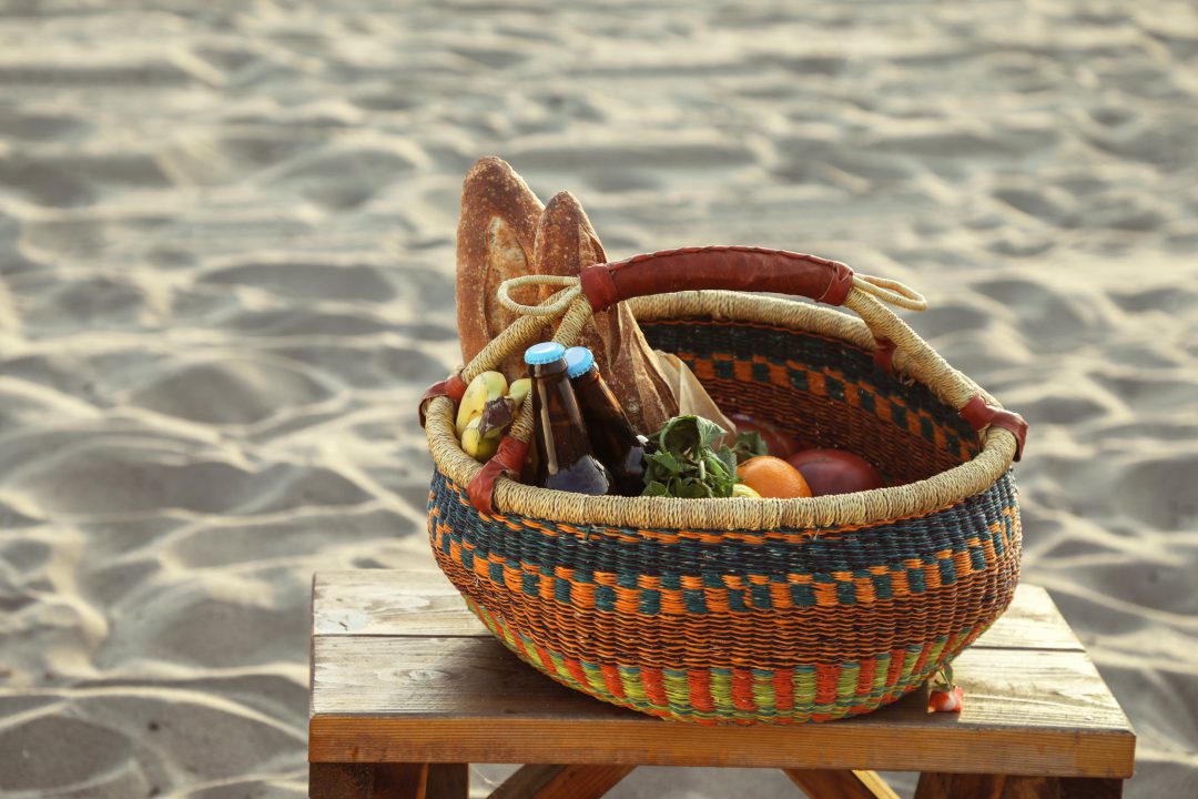 Basket of food and drinks on a wooden table on a sanded beach.