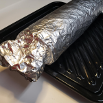Lasagna wrapped in tin foil on a black tray.