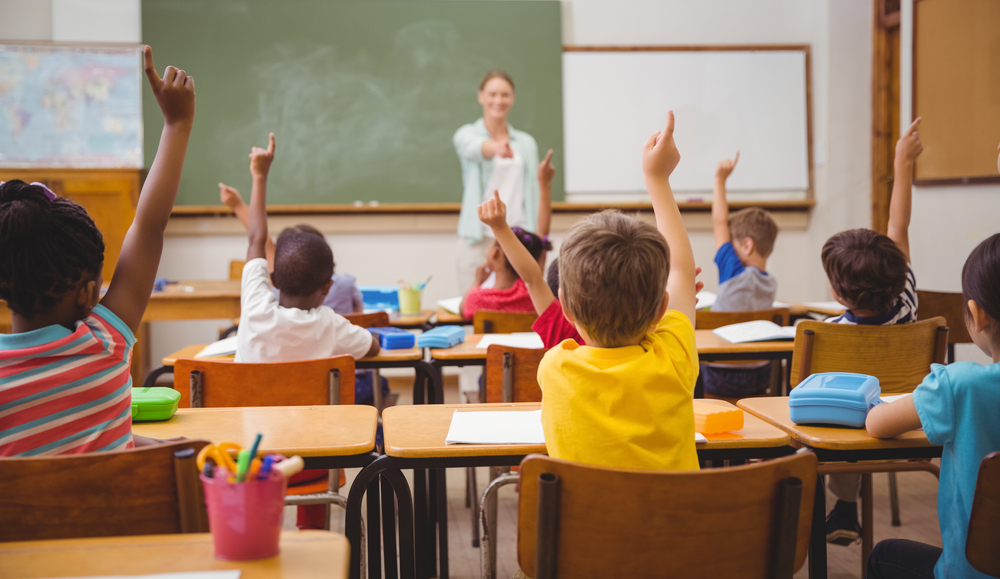 Students raising their hands in a classroom and a teacher pointing at them.