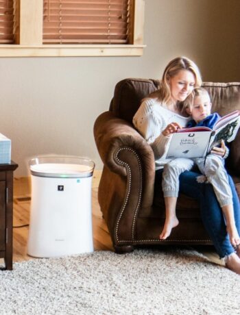 A mom sitting on a chair with her child in her lap reading a book. A Sharp Plasmacluster Ion Air Purifier with True HEPA for Medium Rooms (FPK50UW) is sitting next to the chair.