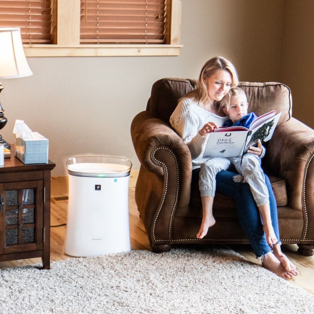 A mom sitting on a chair with her child in her lap reading a book. A Sharp Plasmacluster Ion Air Purifier with True HEPA for Medium Rooms (FPK50UW) is sitting next to the chair.