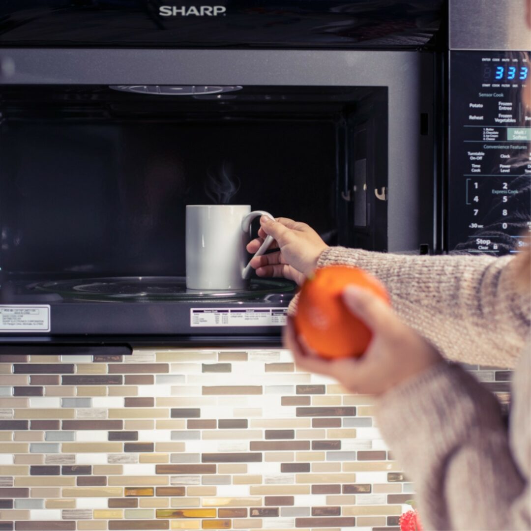 User placing a cup of coffee into a 1.1 cu. ft. 850W Sharp Stainless Steel Convection Over-the-Range Microwave Oven (R1881LSY)