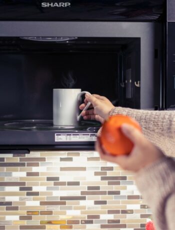 User placing a cup of coffee into a 1.1 cu. ft. 850W Sharp Stainless Steel Convection Over-the-Range Microwave Oven (R1881LSY)