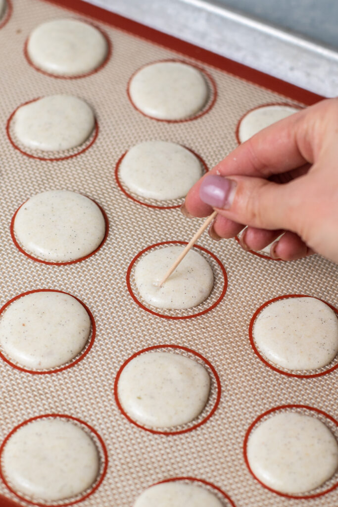 Macarons on a baking sheet being adjusted with a toothpick