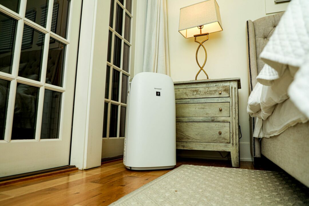 A Sharp KCP110UW Air Purifier sitting beside a nightstand in a bedroom.