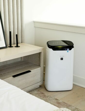 The Sharp Smart Plasmacluster Ion Air Purifier with True HEPA for Extra Large Rooms (FXJ80UW) sitting beside a nightstand in a bedroom.