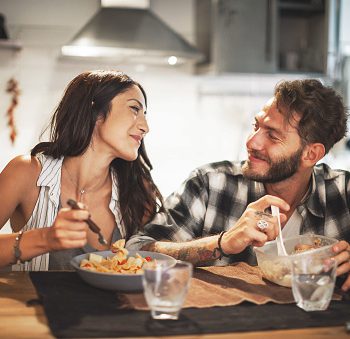 A young couple smiling at one another eating dinner in a kitchen.