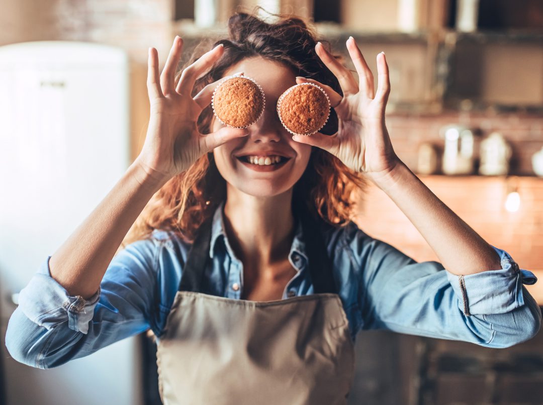 Woman holding two muffins in front of her eyes while smiling in a kitchen.