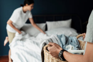 Woman removing bedding from bed while male is holding a laundry basket