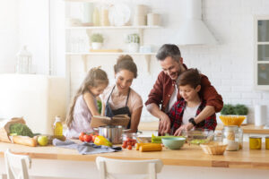 Happy family cooking together on kitchen. Mother and daughter reading recipe to father and son. Dad and boy chopping green vegetable leaf for salad.