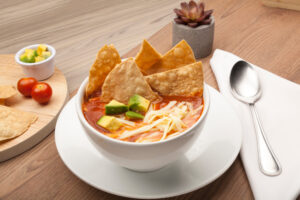 Soup with tortilla chips, avocado, and cheese in a white bowl on a wooden table with spoon