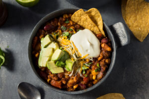 chili with beans, cheese, chips, avocado in a black bowl on a black table 