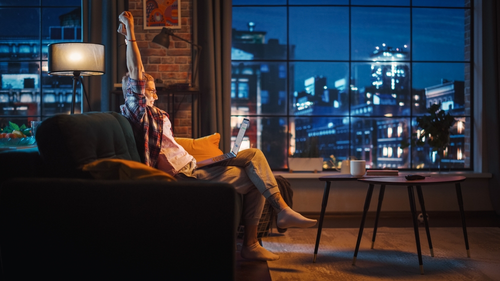 Young Beautiful Woman Sitting on Couch, Using Laptop Computer in Stylish Loft Apartment in the Evening. Creative Female Celebrates Successful News. Urban City View from Big Window.