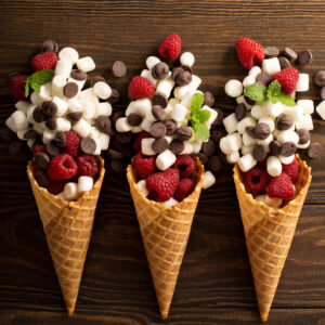 Raspberries, marshmallows, and chocolate chips in an ice cream cone laying on a wooden surface.